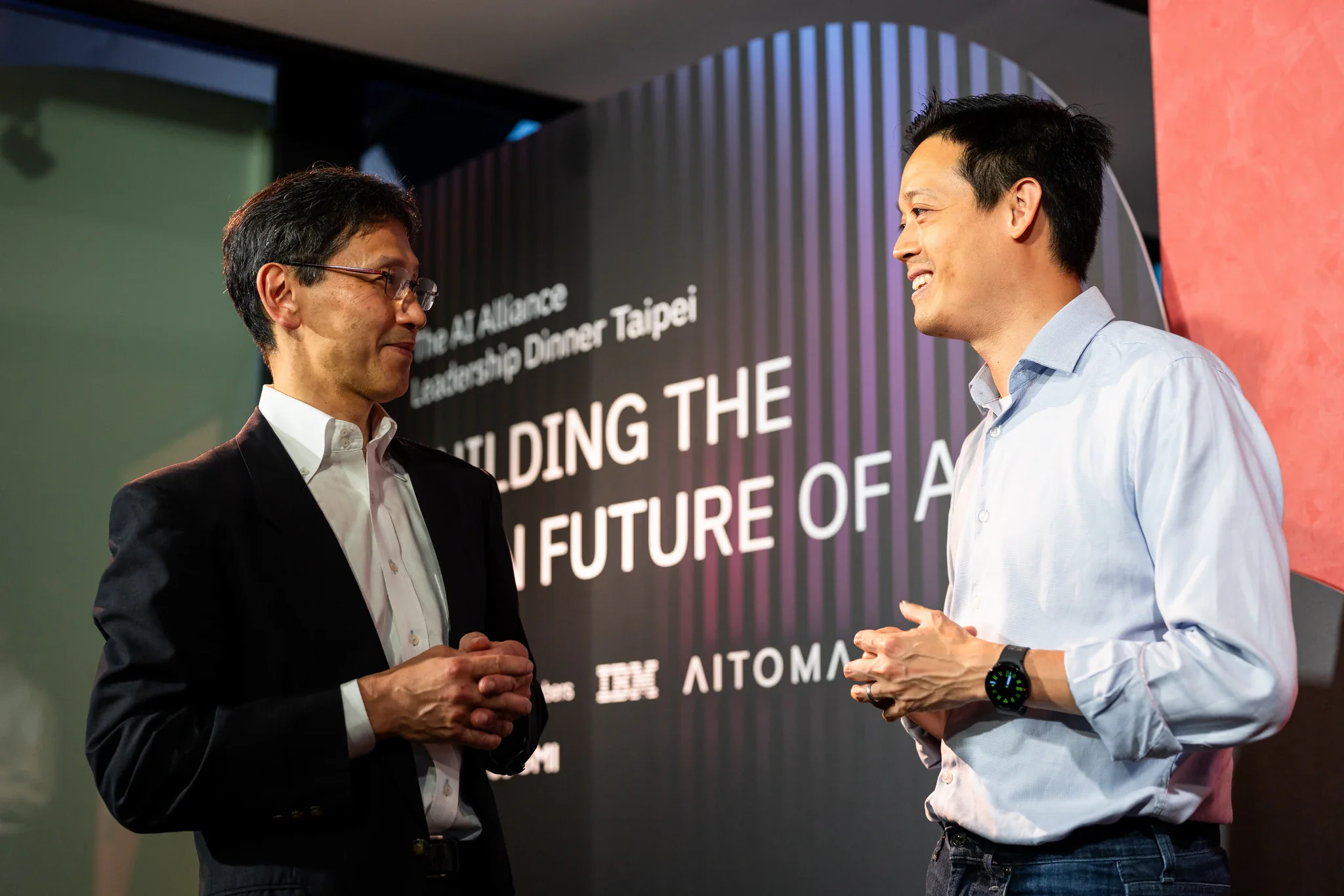 AI Alliance 商務晚宴|品牌牆前的交流 兩位來賓在印有 AI Alliance 活動主題的背板前微笑交談