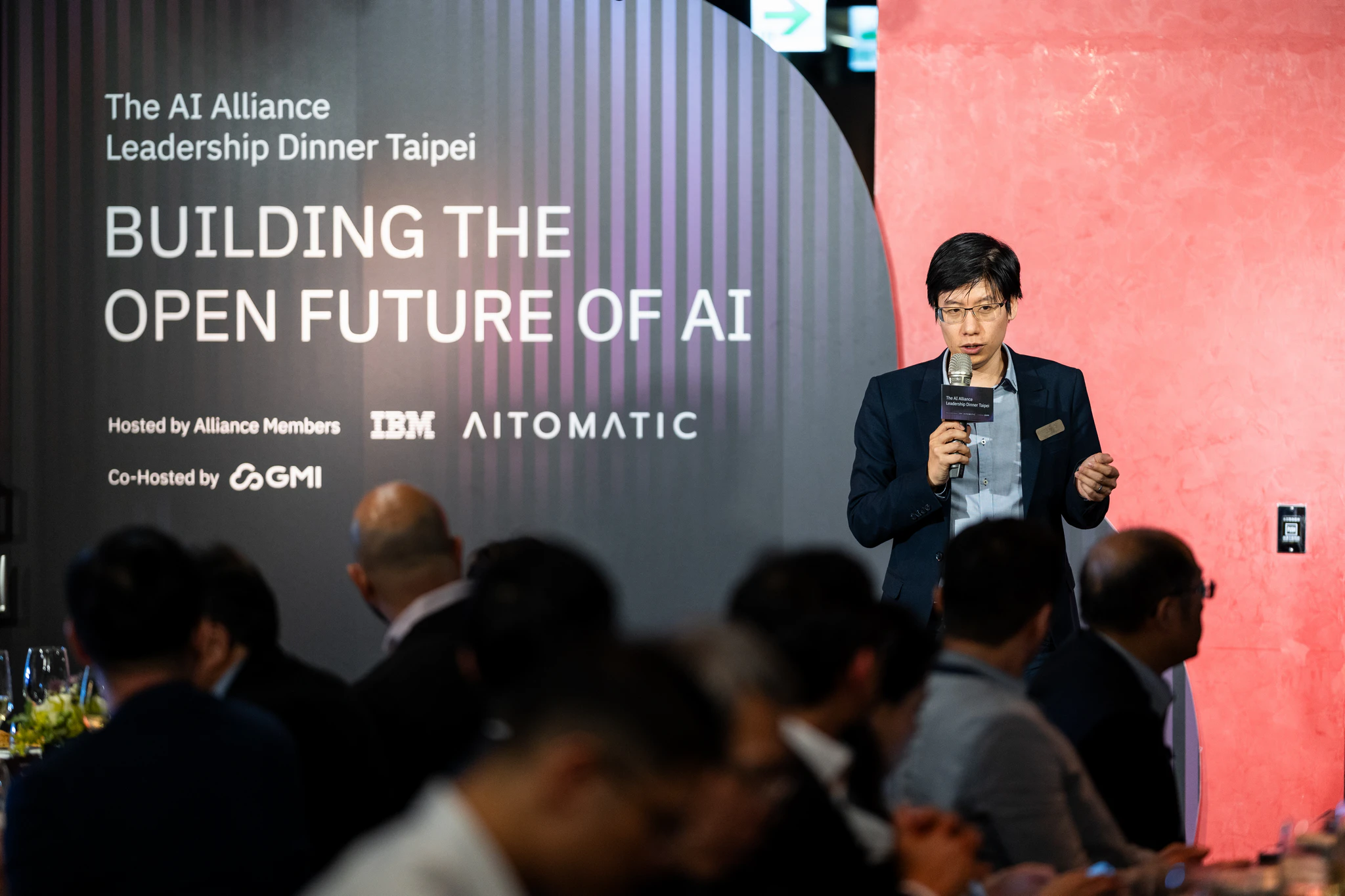 AI Alliance 晚宴攝影|AI 主題演講現場 戴眼鏡的講者在 Building the Open Future of AI 主題背板前持麥克風分享,前景可見賓客