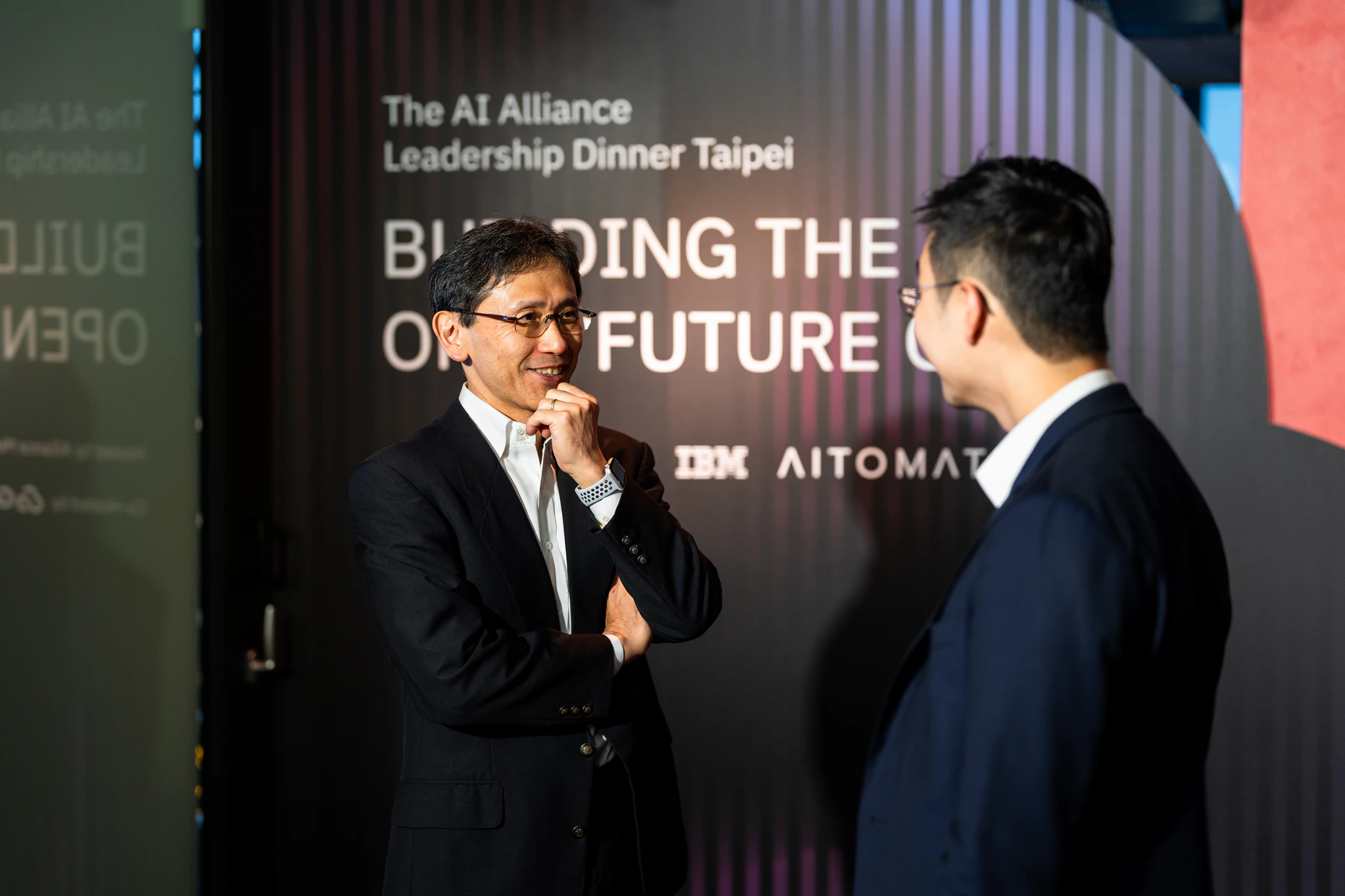 AI Alliance 台北活動攝影|品牌主題牆前交流 兩位穿西裝的來賓在 AI Alliance 主題背板前面對面交談,其中一人微笑聆聽