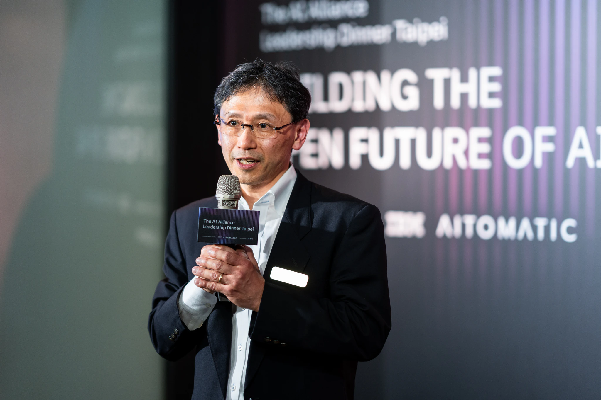 AI Alliance 晚宴攝影|講者與品牌背板 戴眼鏡的男講者手持麥克風,在 AI Alliance 活動主題背板前發言
