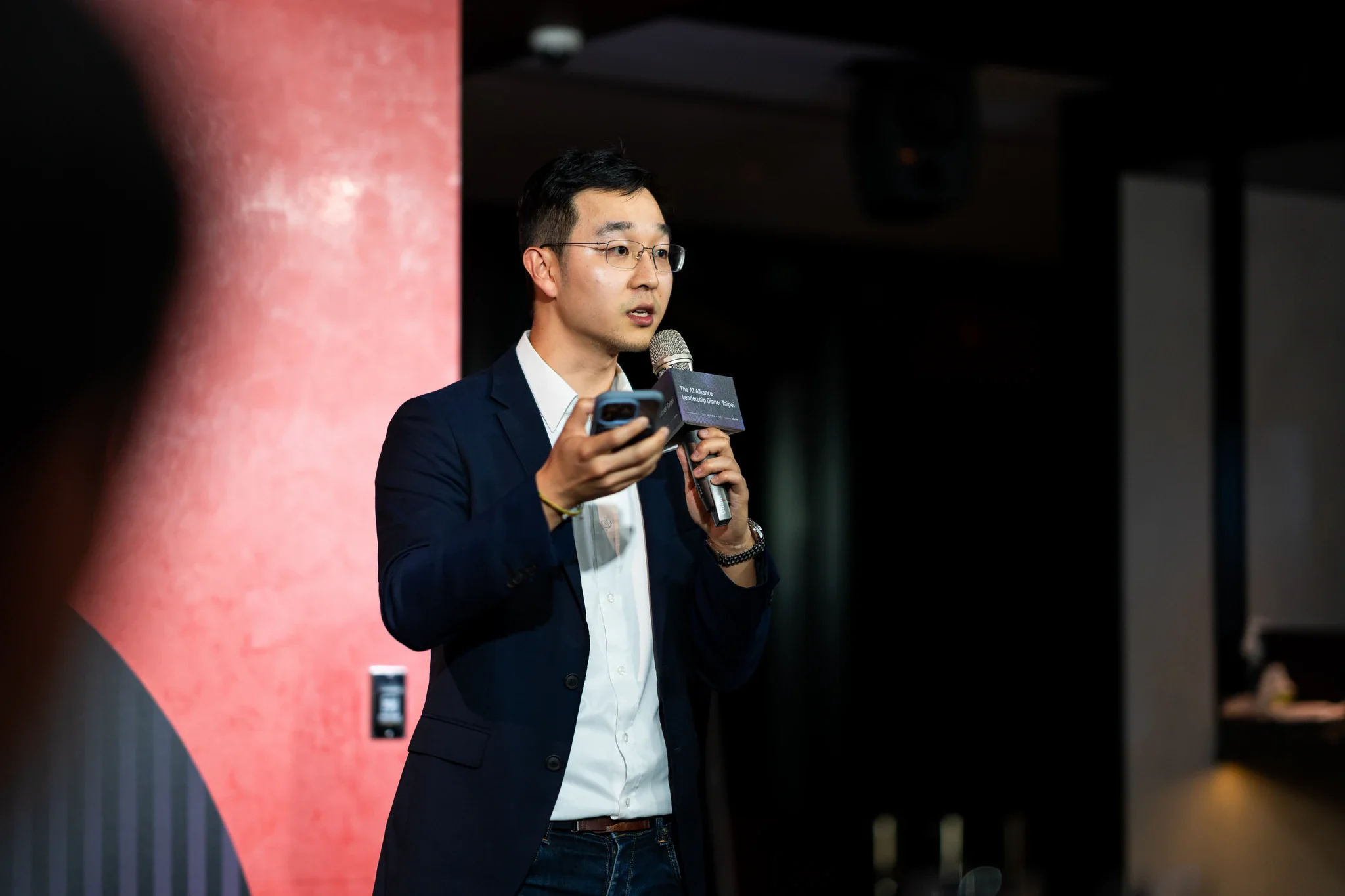 AI Alliance 領袖晚宴|舞台講者特寫 一名西裝講者手持麥克風在深色背景前發言