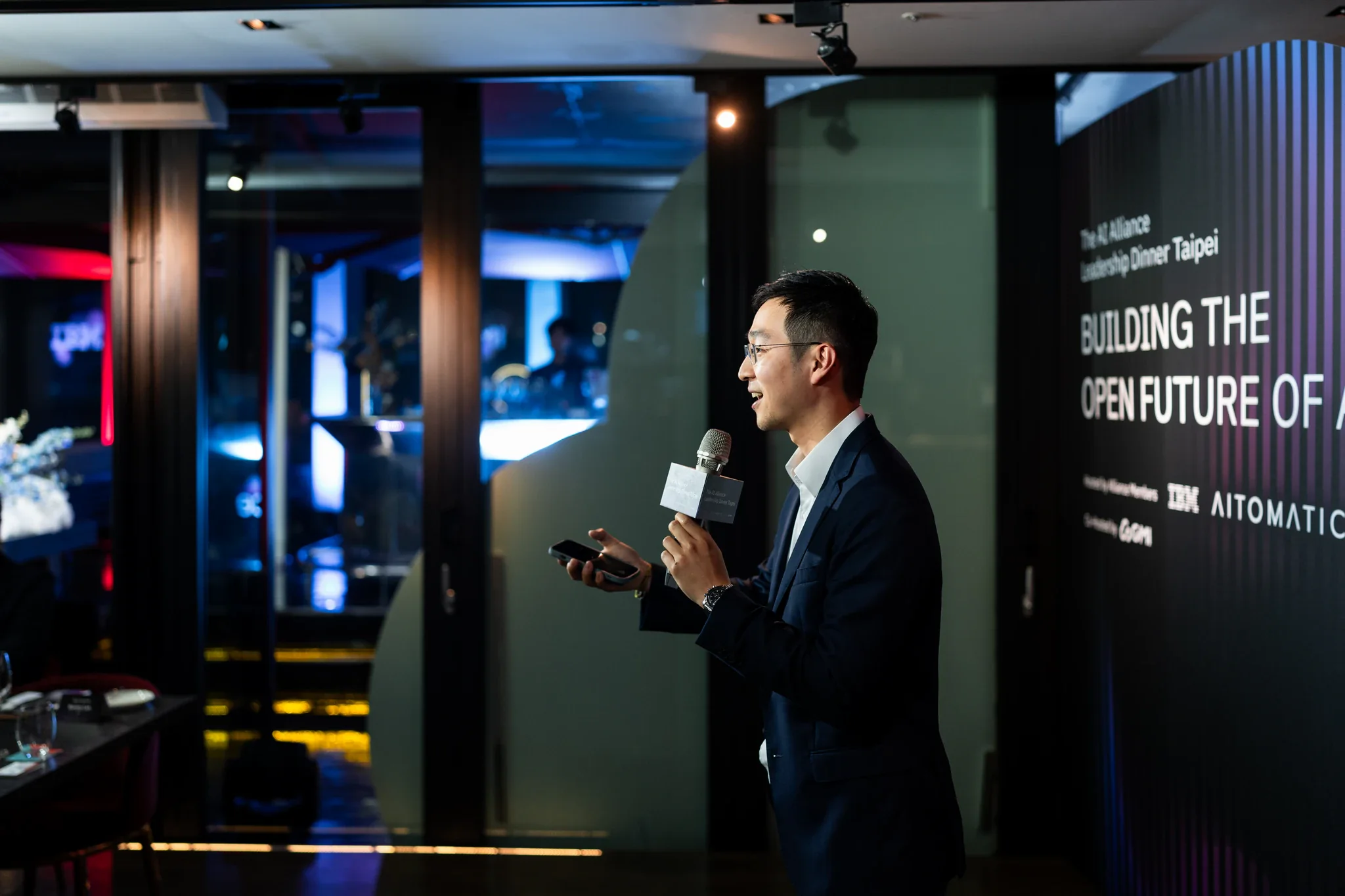 AI Alliance 領袖晚宴攝影|講者致詞 演講者站在活動主題牆旁,以麥克風向來賓說明