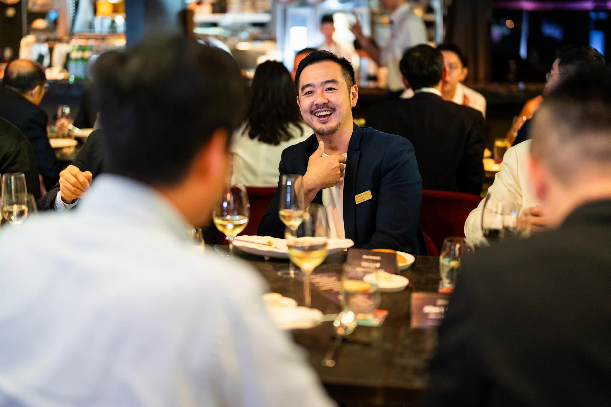 AI Alliance 商務晚宴|餐桌間的自然交流 穿深色外套的男士在晚宴餐桌間微笑交談,前景可見其他賓客與酒杯