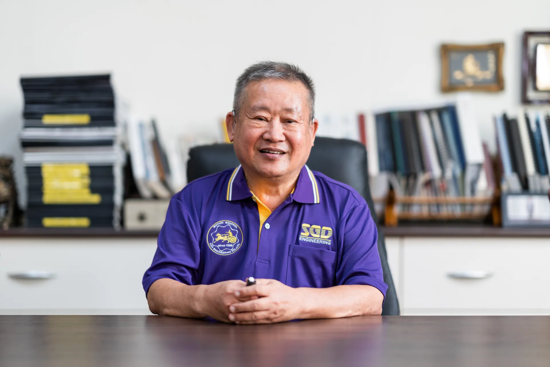 協聚德企業人像攝影|自然辦公室肖像 一名身穿協聚德紫色制服的男性員工坐在辦公室桌前,面向鏡頭微笑