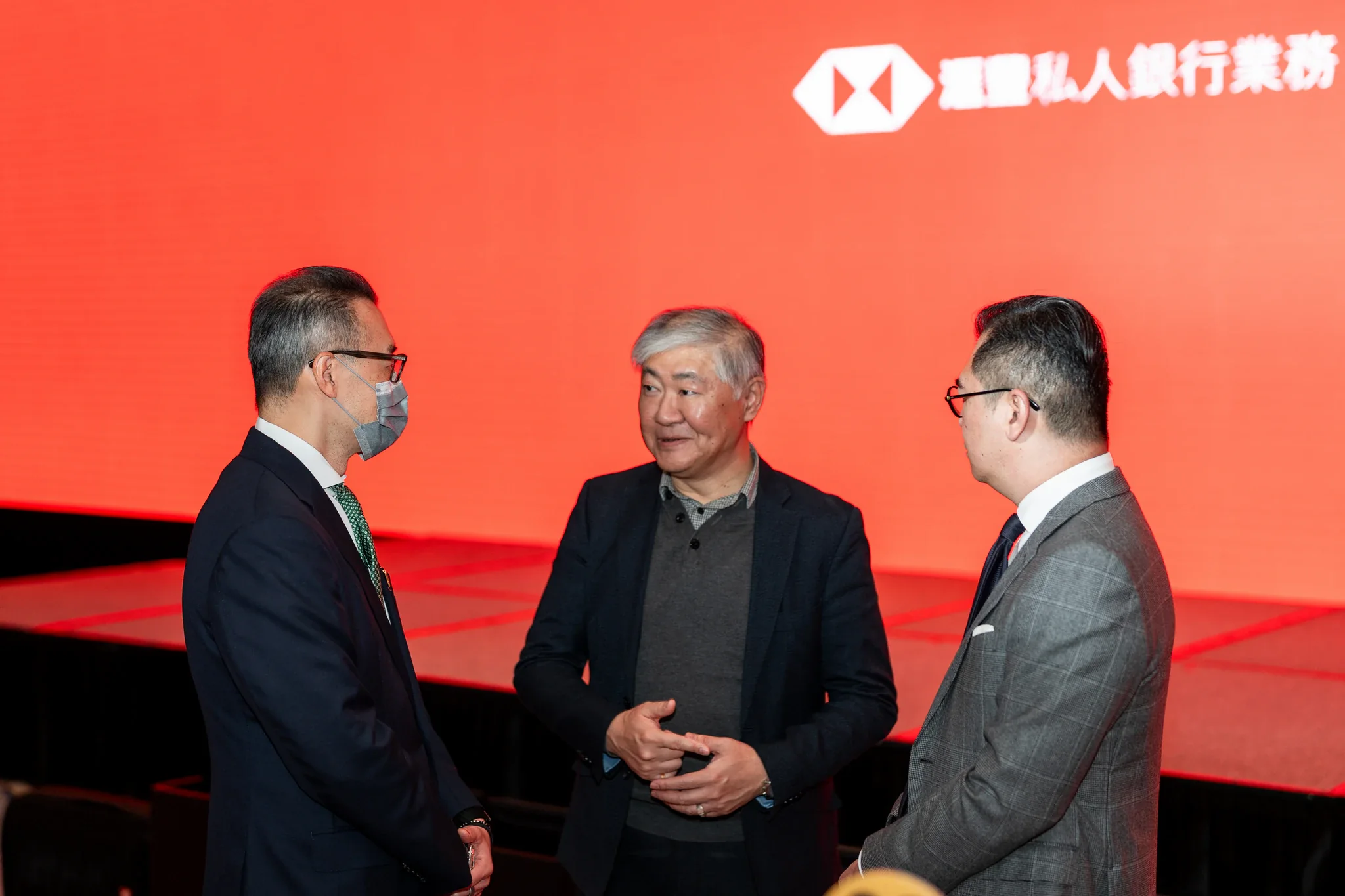 滙豐銀行活動攝影|品牌背板前的交流 三位來賓在帶有滙豐標誌的紅色活動背板前交談
