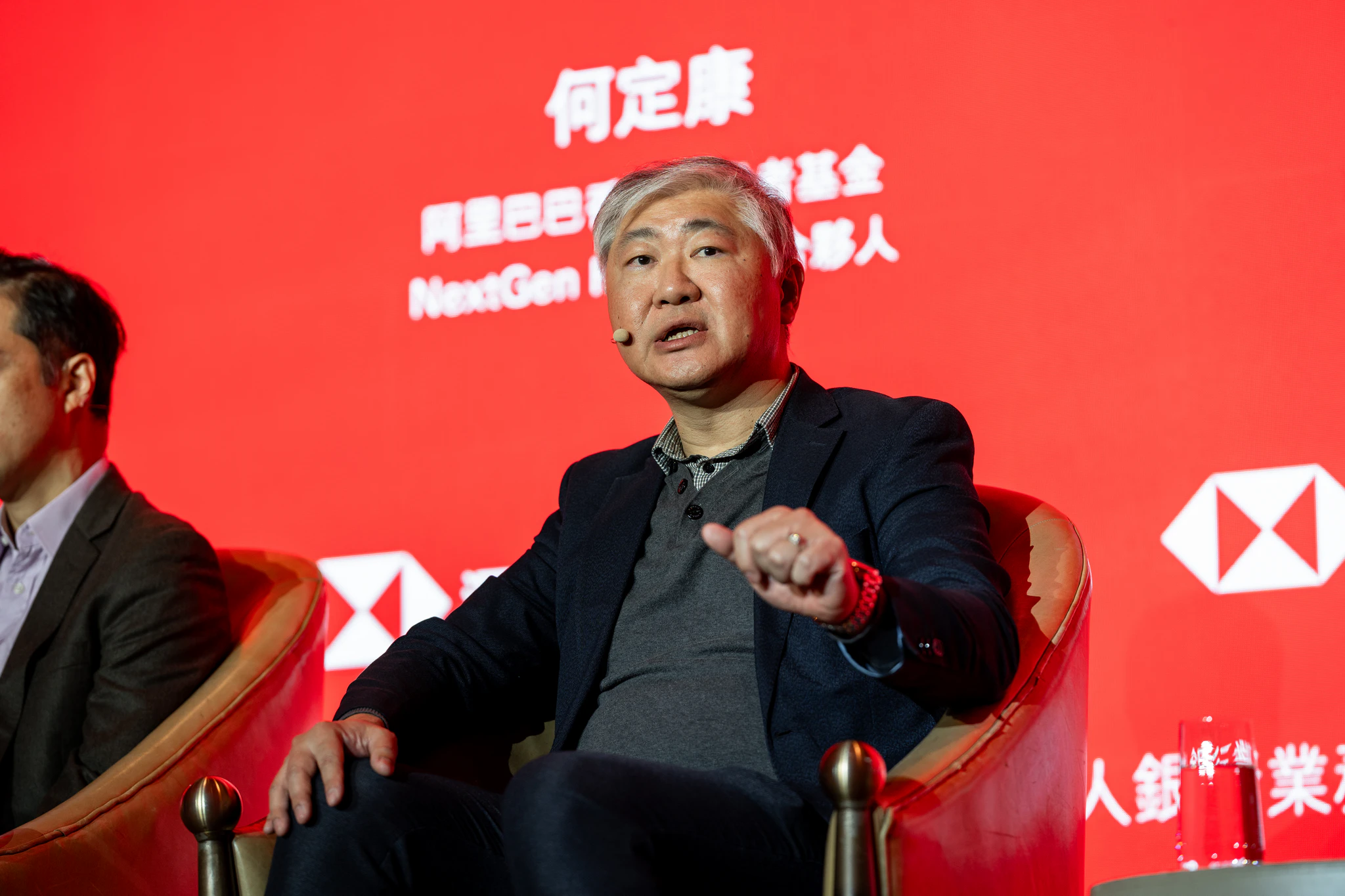 滙豐銀行投資展望|座談講者特寫 銀髮男與談者坐在滙豐紅色活動舞台上發言,後方可見講者姓名與品牌視覺
