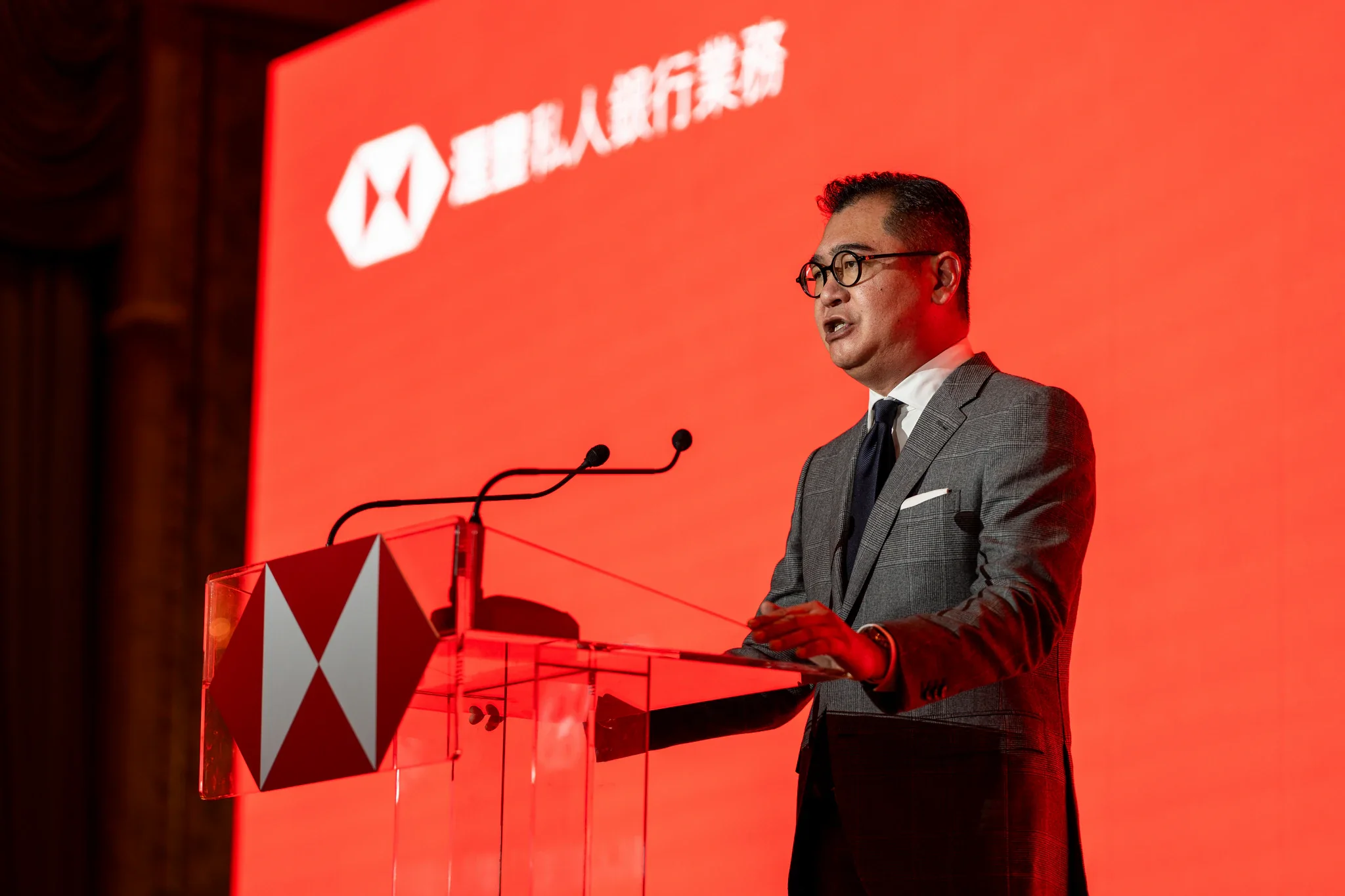 滙豐銀行 2026 投資展望|講台發言 一名穿灰色西裝的男講者站在印有滙豐標誌的透明講台前發言